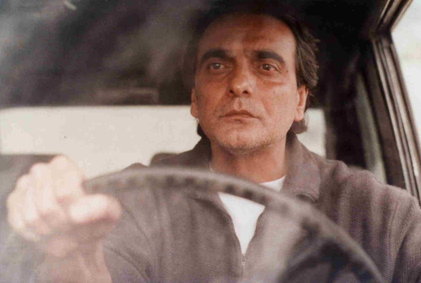 A middle-aged Iranian man with sad, sunken eyes stares out the windshield of his car with his hand on the steering wheel.