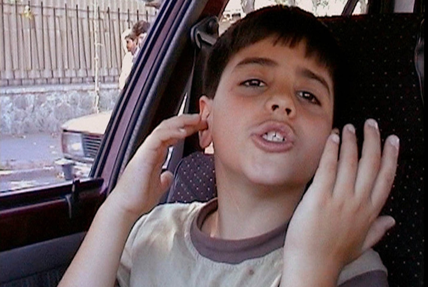 A young Iranian boy sitting in the passenger seat of his car, throwing his hands up with a frustrated expression on his face.