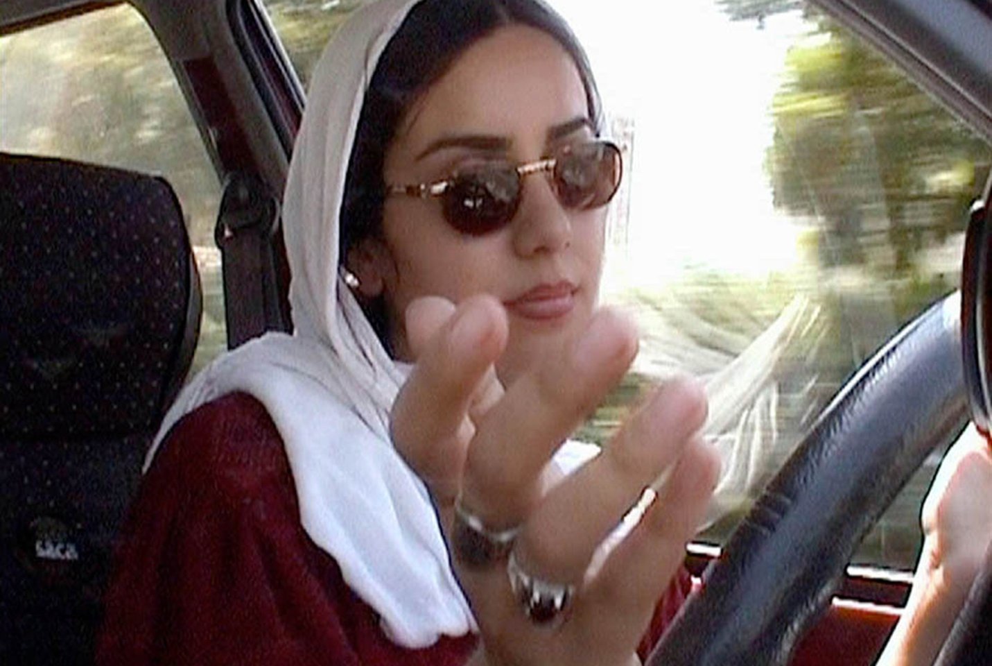 An Iranian woman wearing a white headscarf and sunglasses sitting in the driver's seat of a car, gesticulating at the camera with one hand.