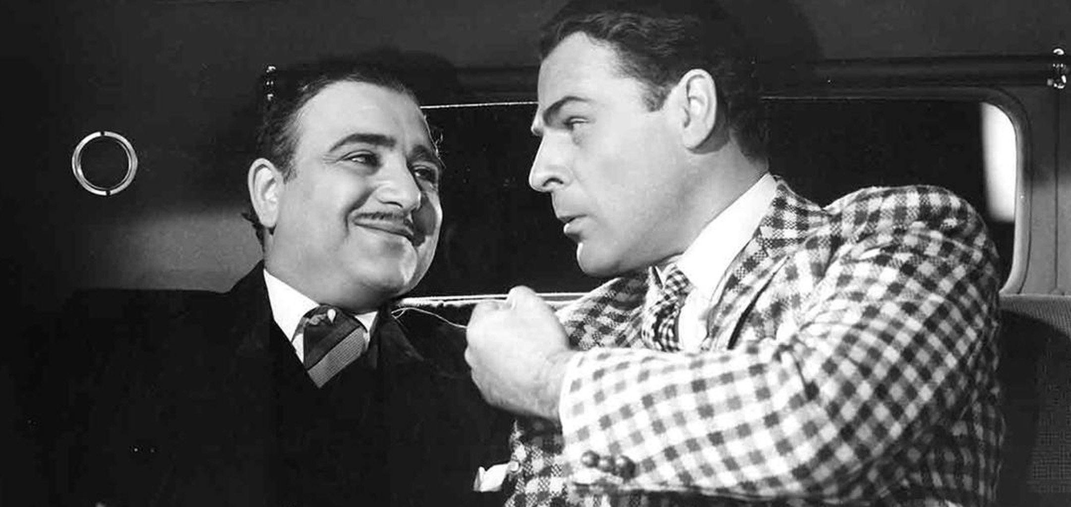 Akim Tamiroff and Brian Donlevy in the back of a cab in a black and white still from the film 'The Great McGinty' (1940). Tamiroff (left) is wearing a dark suit and is smiling at Donlevy, who is wearing a chequered suit and gesticulating with his fist.