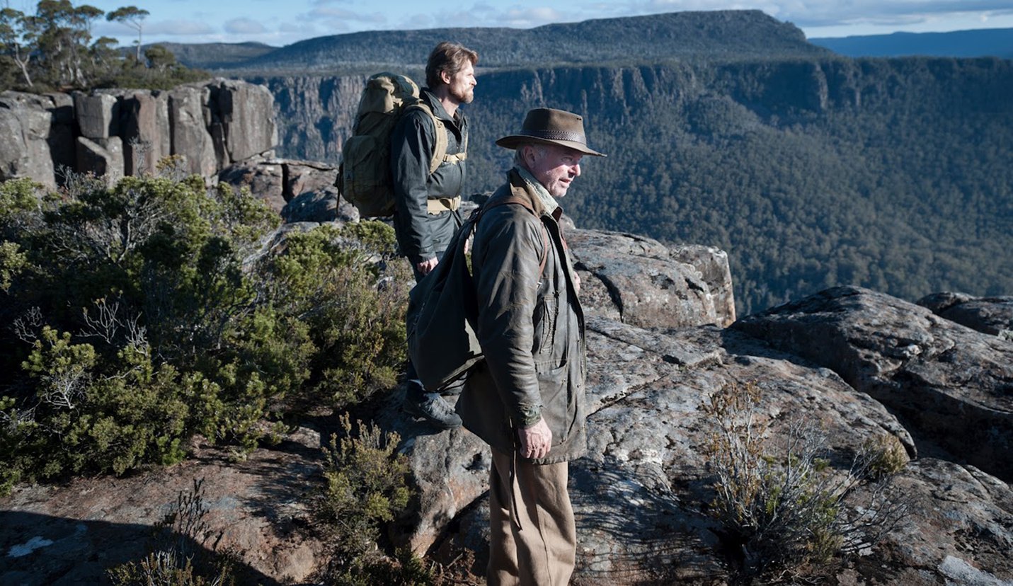 Willem Dafoe as Martin David (background) and Sam Neill as Jack Mindy (foreground) in 'The Hunter' (2011). They are both standing on a rocky outcrop overlooking a spectacular valley in the Tasmanian wilderness in daylight.
