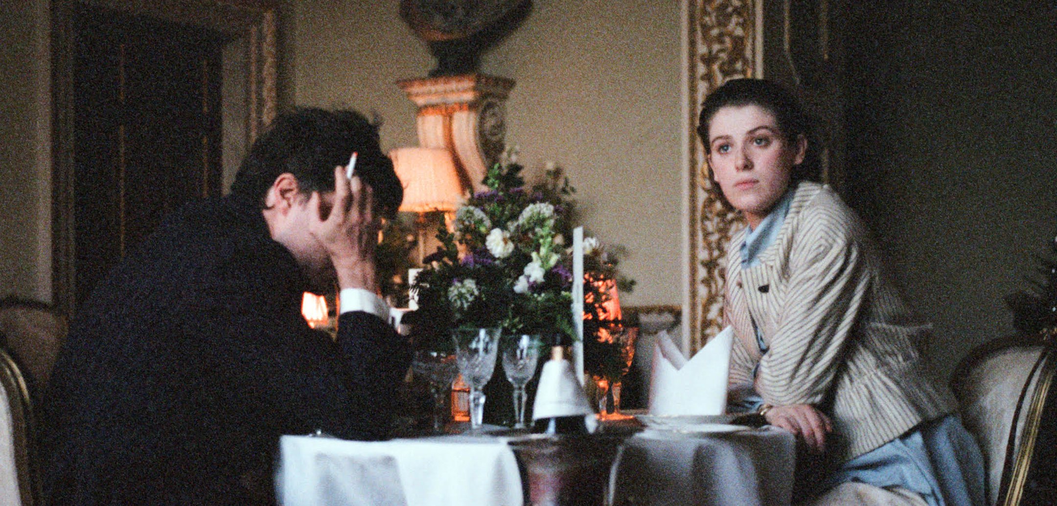 A still from the film 'The Souvenir' (2019). Tom Burke and Honor Swinton Byrne in sit at a tale in an upmarket restaurant. Burke's face is covered by his hand, in which is a cigarette, while Swinton Byrne looks off into the distance.