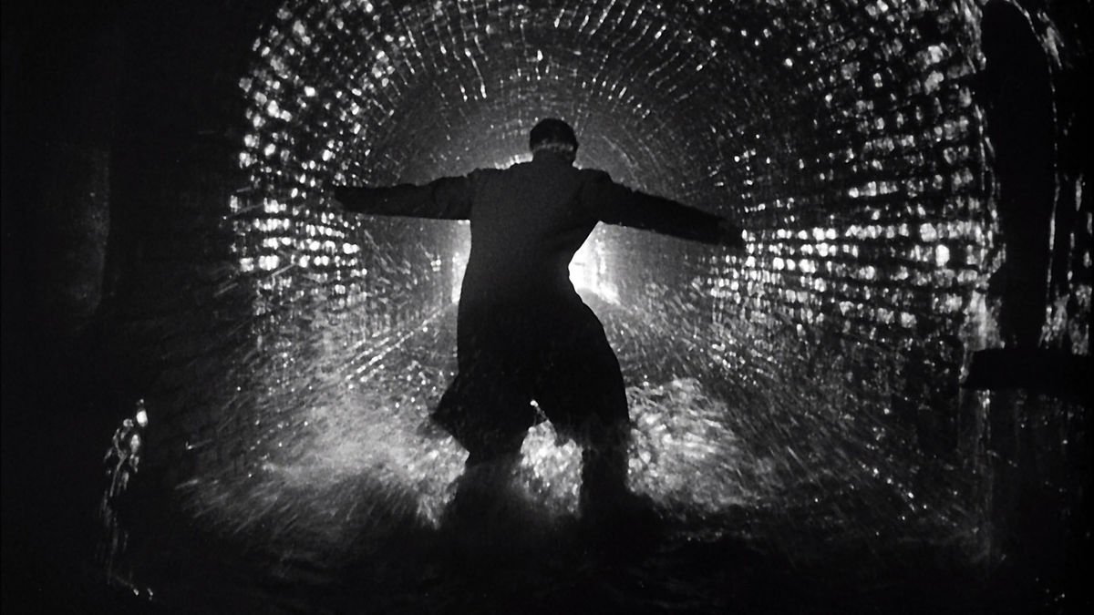 An image of a man in a black coat in a sewer with his arms outstretched, looking down towards the tunnel of light, the wet bricks glistening like fireflies.