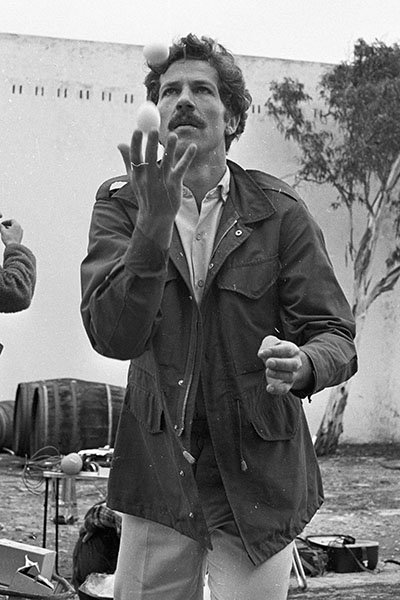 A three-quarter length portrait photo of Werner Herzog, in black and white from the 1970s. He is wearing a dark jacket, juggling 2 eggs and has a look of concentration on his face.