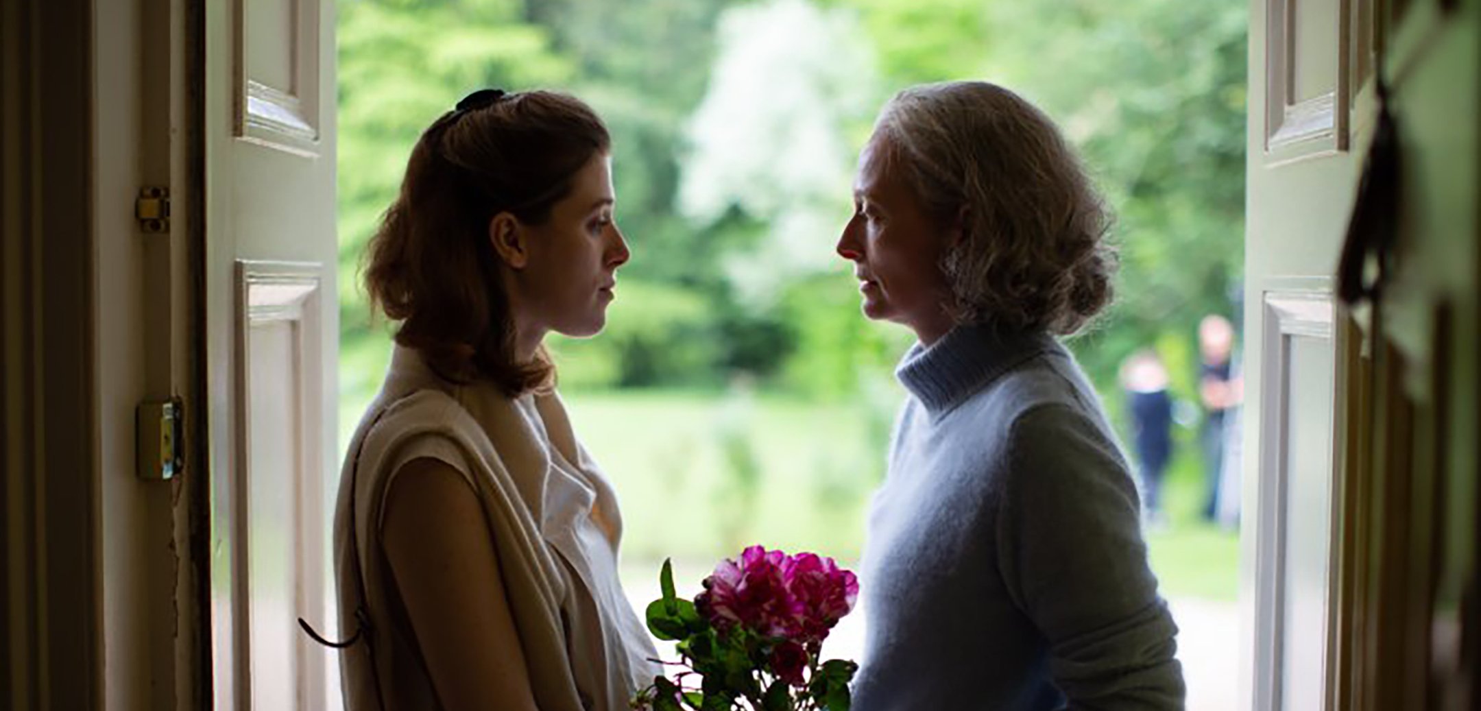 A young woman (left) and an older woman (right) face each other in a doorway in partial silhouette with a bouquet of pink flowers between them.