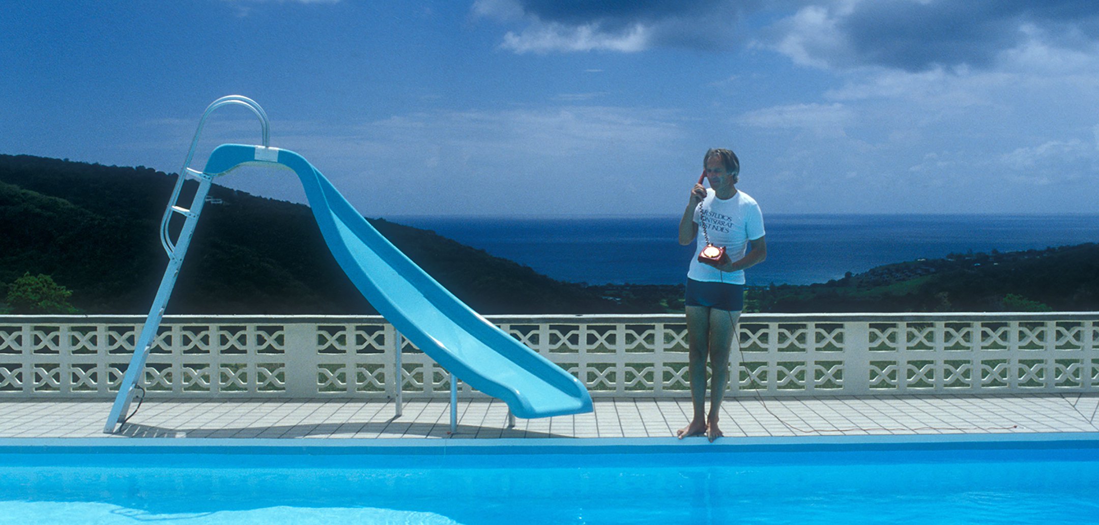 Still from Under the Volcano (2021) - George Martin by a blue pool in an island setting