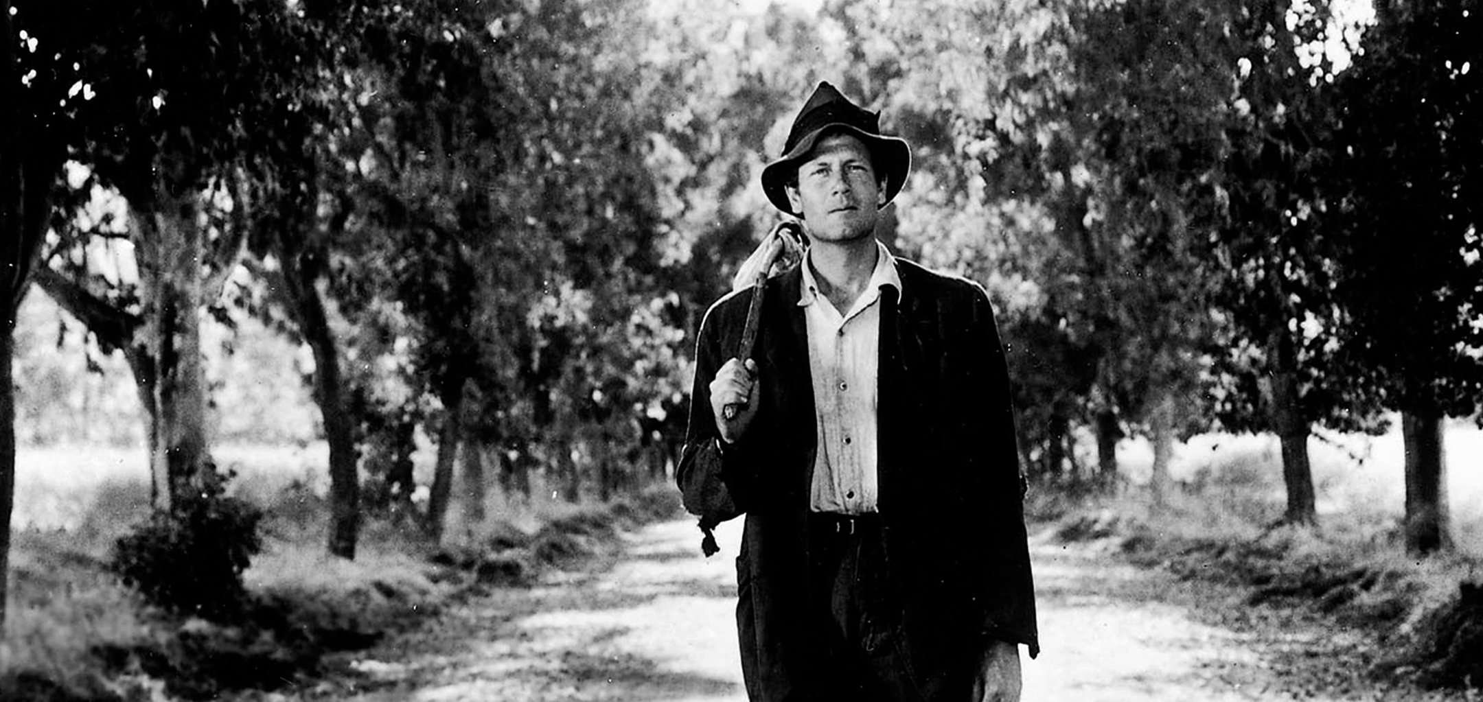 Joel McCrea in a black and white still from the 1941 film 'Sullivan's Travel's' directed by Preston Sturges. He is wearing a suit and a wide-brimmed hat and is carrying a bindle over his shoulder as he walks down a tree lined country road.