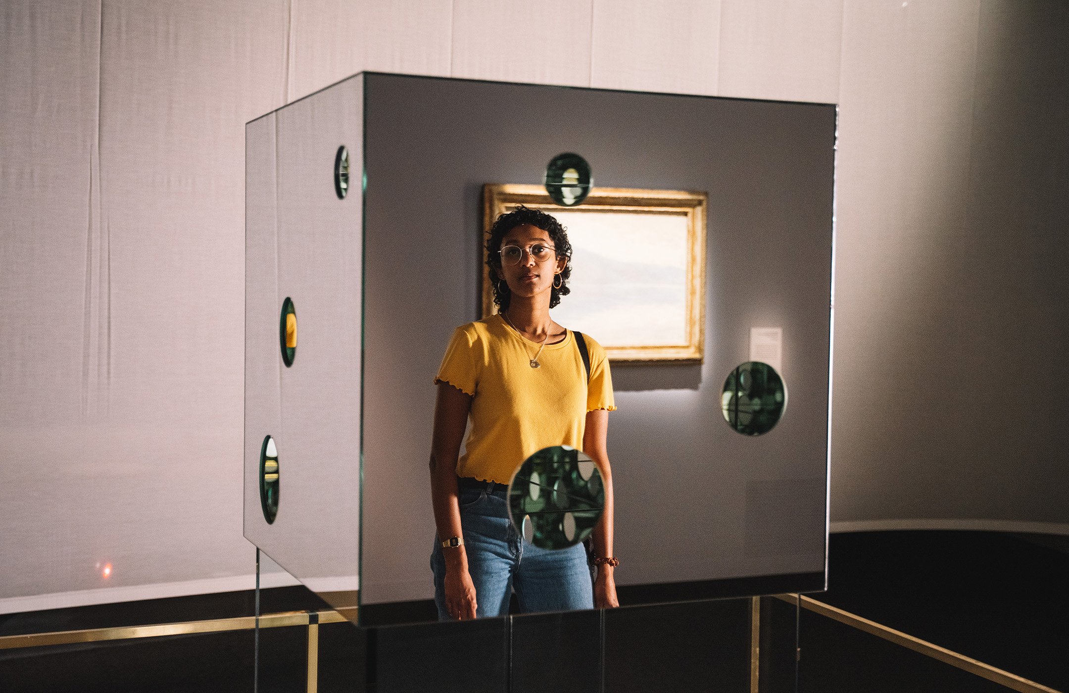 A young woman peers into a glass cube with holes in it (Yayoi Kusama's artwork The Passing Winter). She is wearing glasses and a bright yellow top, and has a dark complexion and short curly hair.