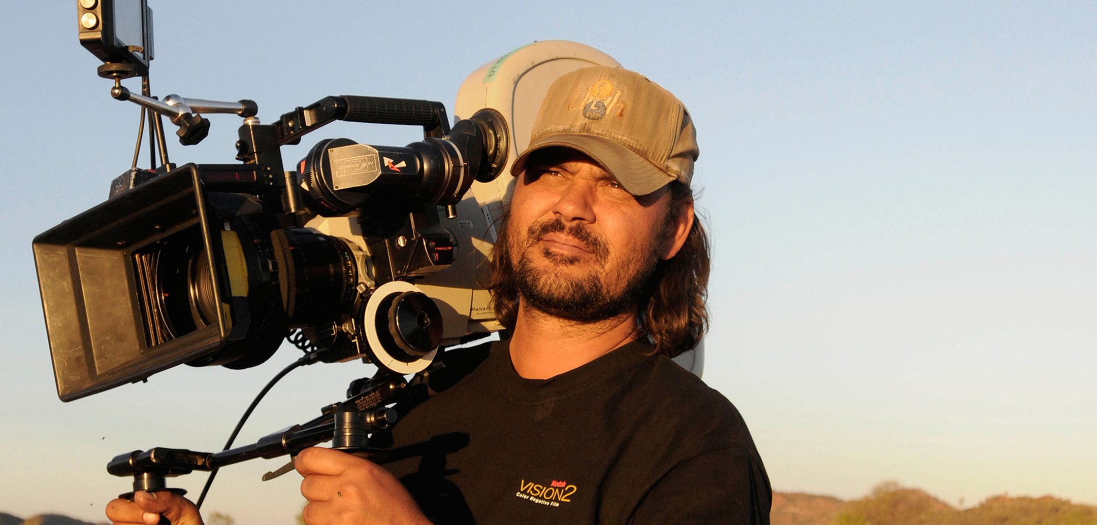 Filmmaker Warwick Thornton wearing a cap and holding a large film camera on his shoulder.
