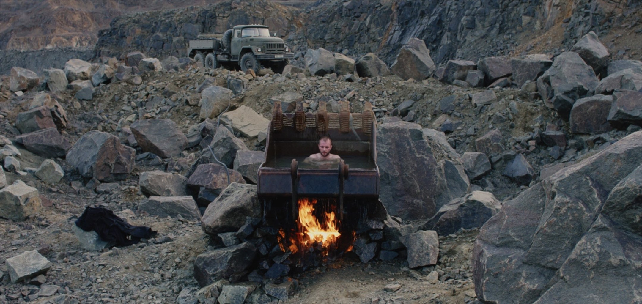 A barren, rocky quarry in Ukraine with a man sitting in water collected in the digging scoop of mining machinery, which is being heated by fire underneath.