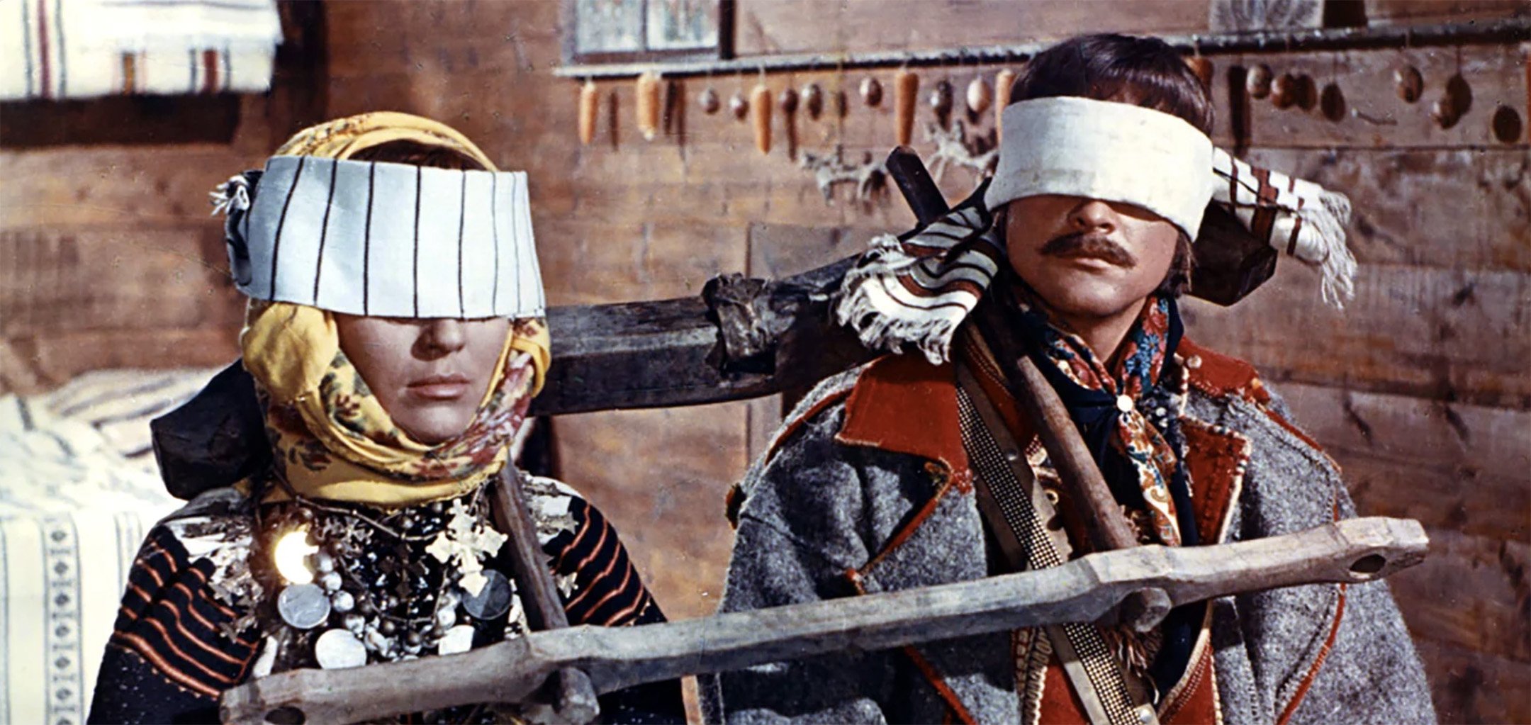 A woman and a man in traditional Ukrainian clothes blindfolded and carrying a wooden contraption on their shoulders.